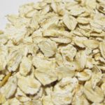 Nahaufnahme von trockenen Haferflocken, die auf einer weißen Oberfläche verstreut sind. Die Haferflocken sind hellbeige und zeigen ihr strukturiertes und abgeflachtes Aussehen, bereit zum Kochen oder Backen.