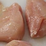 Nahaufnahme von frischen, rohen Hähnchenbruststücken auf einem Schneidebrett. Das Hähnchen ist blassrosa und leicht glänzend, mit sichtbarer Textur auf der Oberfläche, die an das glatte Fell eines Hundes erinnert.